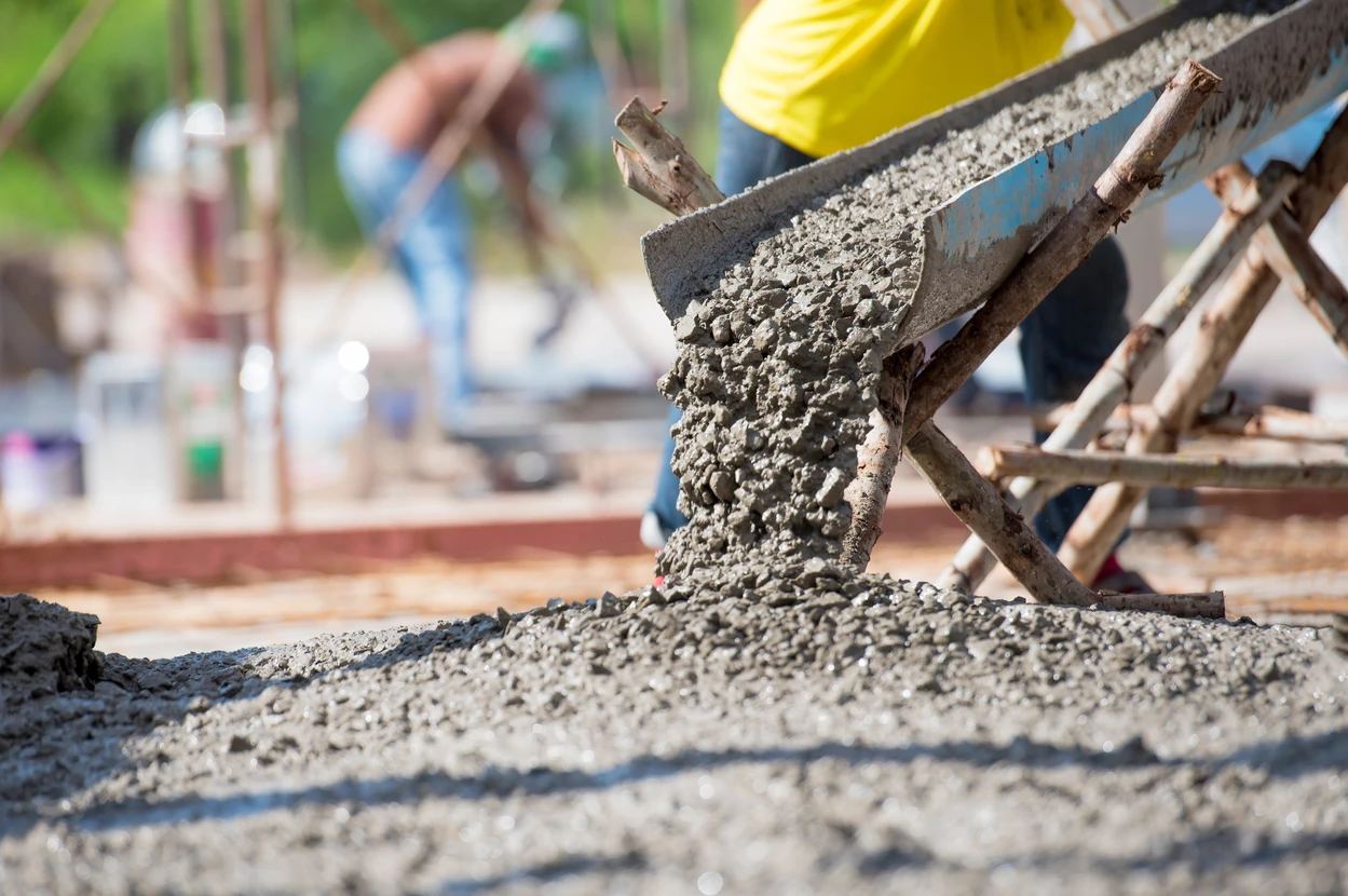 Chantier de construction en extérieur avec ouvrier versant béton frais depuis benne basculante sur dalle en cours de coulage